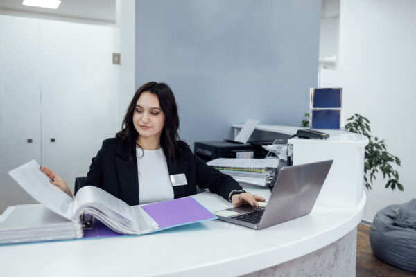 Office female worker in black jacket and white shirt works with catalog of papers and laptop. Young pretty business woman enters information into computer. Stacks of papers and printer on background.